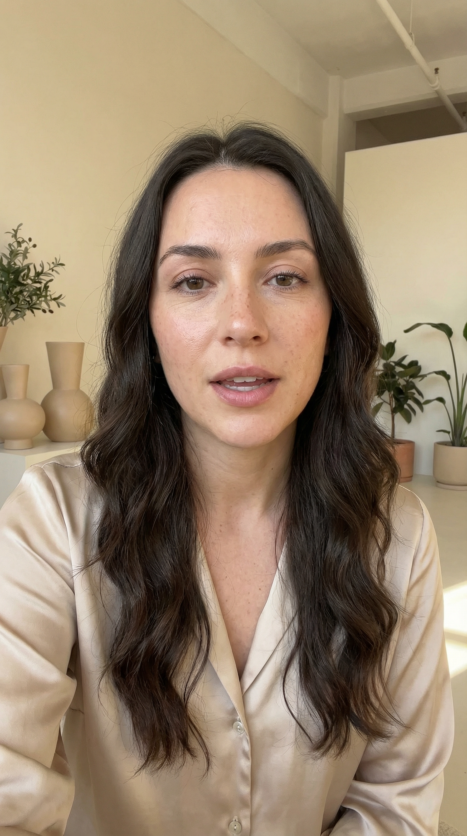 Photoreal portrait of a fictional talk-to-camera creator in a champagne blouse inside a soft neutral studio apartment.